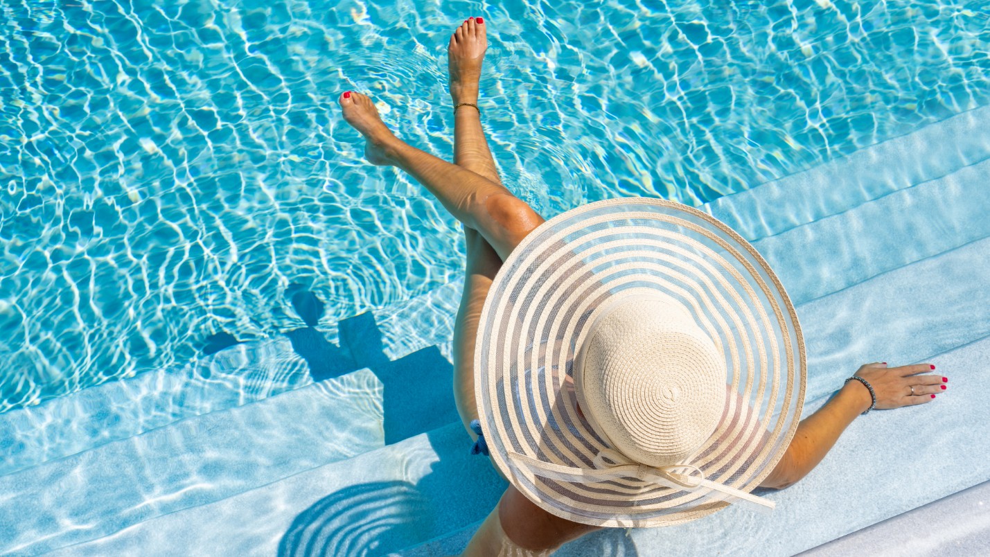 A woman in a large sunhat sits on the steps of a crystal clear pool.