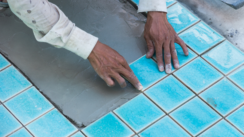 A pool builder carefully places blue tiles onto wet cement.
