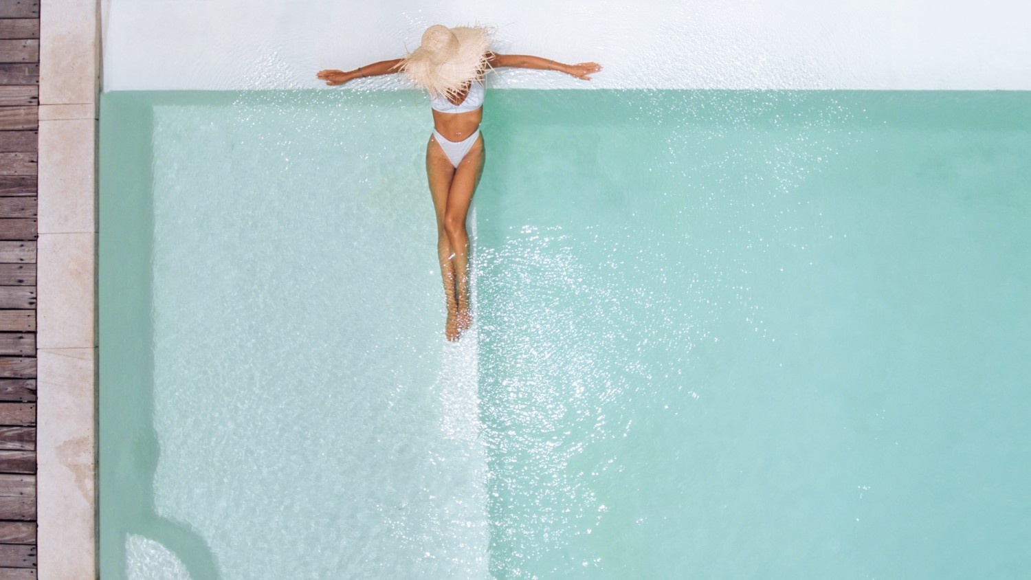 A woman in a sun hat lounges on the tanning ledge in her large luxury pool.