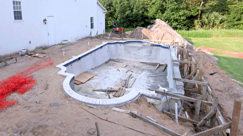 A pool under construction in a backyard next to a white house and lawn.