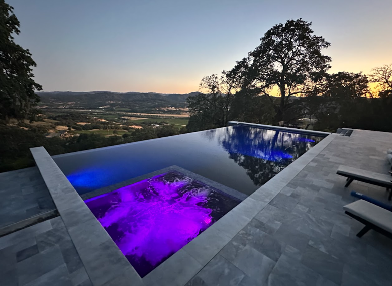 Deep blue infinity pool with purple-lit spa, gray stone tile deck, and tree reflections overlooking rolling hills at sunset.