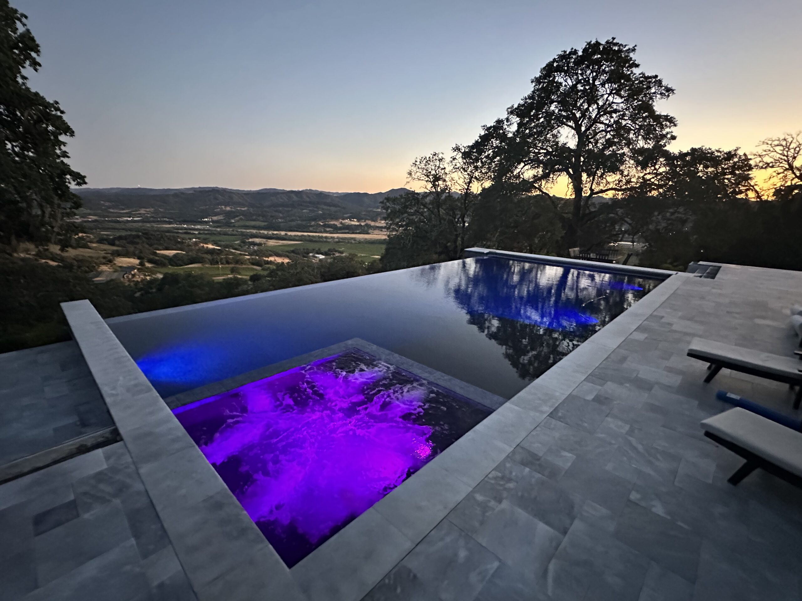 Deep blue infinity pool with purple-lit spa, gray stone tile deck, and tree reflections overlooking rolling hills at sunset.