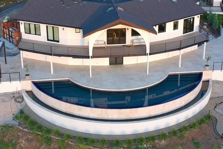 Aerial view of a white hillside home with a dark brown roof, curved black railings, and a crescent-shaped deep blue infinity pool.